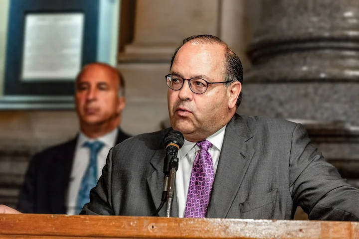Allan Domb speaking at a podium in Philadelphia City Hall, dressed in a gray suit and purple tie, with another suited man in the background.