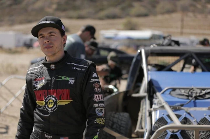 Brad Deberti standing beside a custom-built off-road truck, wearing racing gear and smiling at the camera.