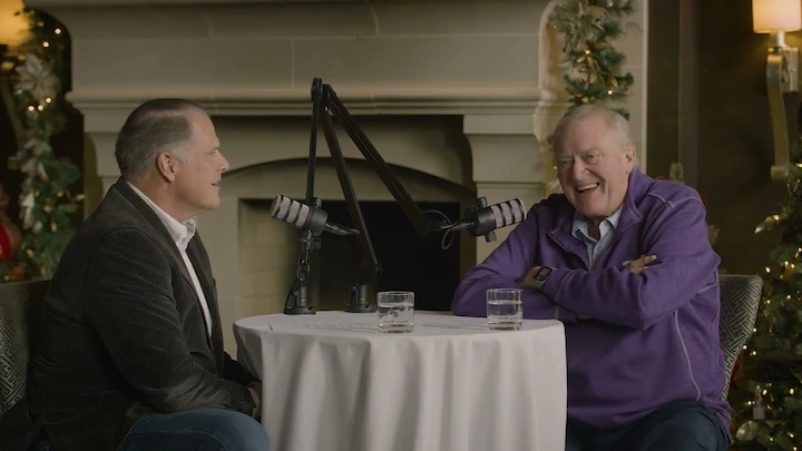 Two older men recording a podcast together at a cozy table, sharing laughter in a warmly decorated living room with holiday lights.
